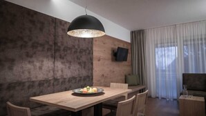 Un espace repas moderne avec une table en bois, des chaises, un luminaire suspendu noir, un téléviseur fixé au mur et une vue sur l’extérieur par une fenêtre agrémentée de rideaux.