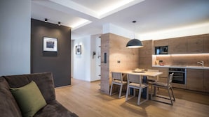 A modern kitchen with wooden cabinets, a dining area with a table and chairs, and a living room with a sofa and a green pillow.