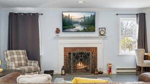 A living room with a fireplace, a plaid armchair, a sofa, and a television mounted above the fireplace.