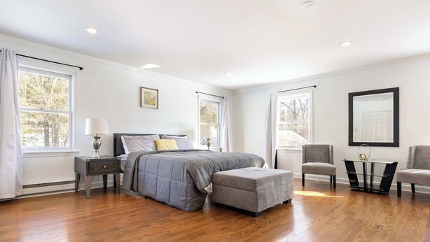 A bedroom with a bed, nightstand, chair, ottoman, and a table with a lamp.