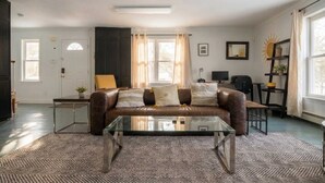 A living room with a brown leather sofa, a glass coffee table, a dark wooden cabinet, a small side table, and a large window with curtains.