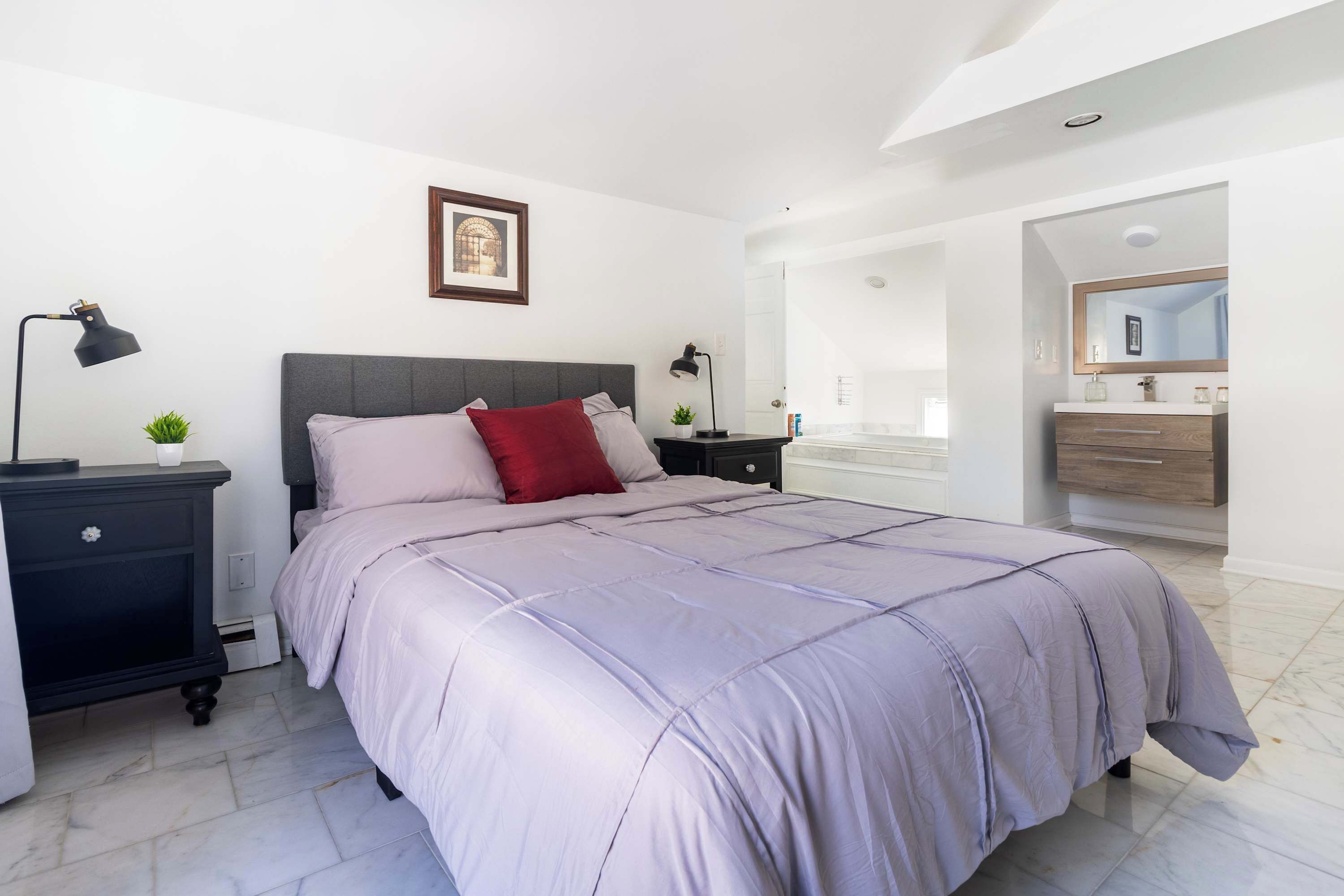 A modern bedroom with a large bed, bedside tables, a nightstand, a framed picture, and a bathroom with a mirror and vanity.