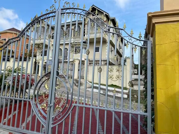 Uma entrada com portão e detalhes metálicos elaborados, um edifício com vários andares e varandas, e uma parede amarela à direita.