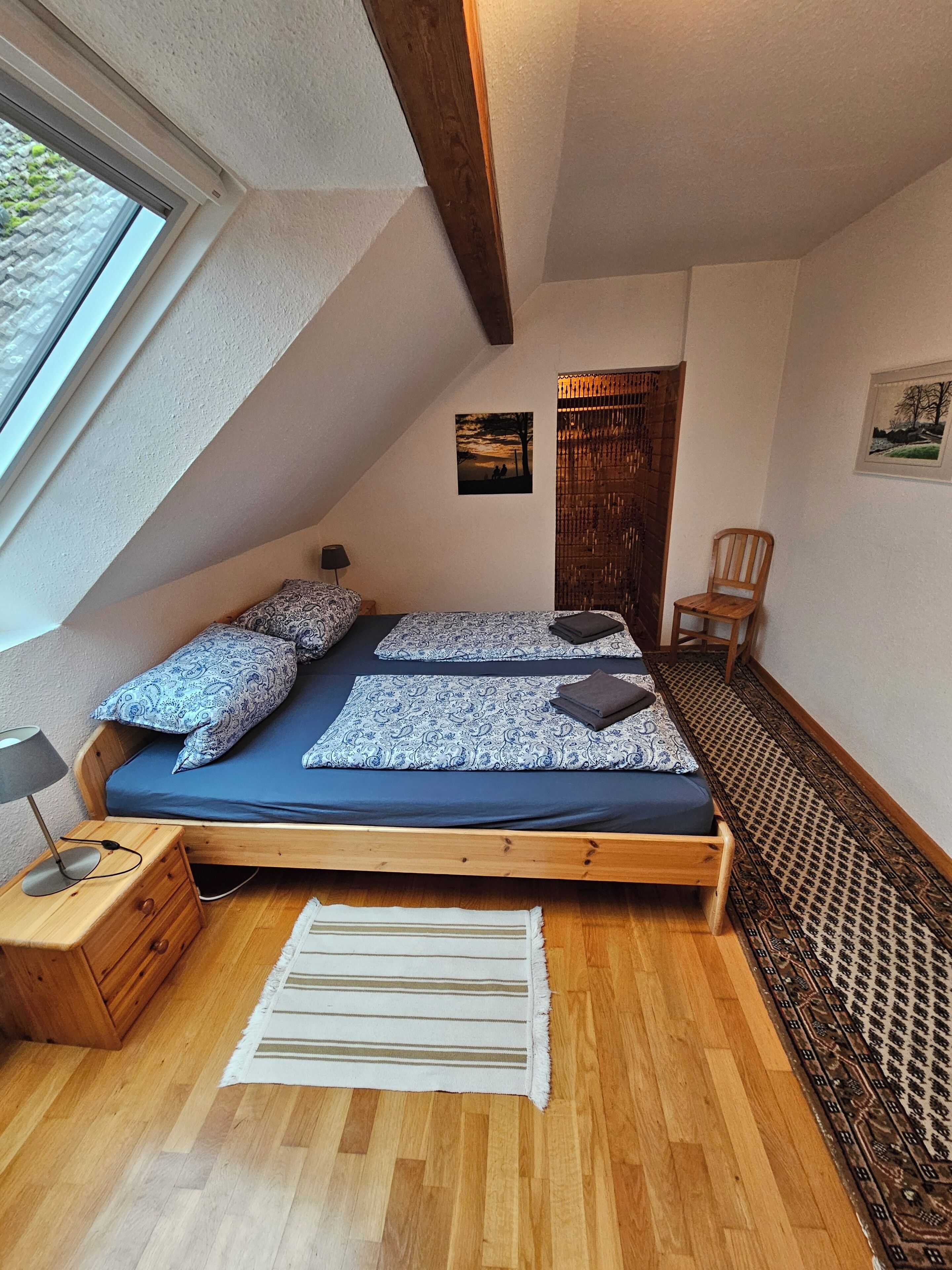 Un dormitorio con techo inclinado, cama con ropa de cama estampada, mesita de noche de madera, una silla y vistas al exterior.