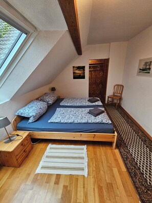Une chambre avec un plafond en pente, un lit avec une literie à motifs, une table de chevet en bois, une chaise et une vue sur l’extérieur.