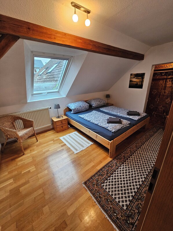 Quarto com teto inclinado, claraboia, cama com roupa de cama estampada, piso de madeira, uma cadeira, mesa de cabeceira e tapete.
