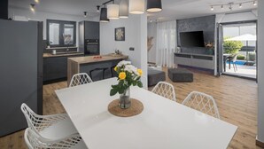 Una cuina moderna amb una illa central, una zona de menjador amb taula i cadires blanques, i una sala d'estar amb televisió i seients.