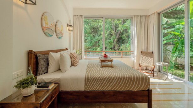 Une chambre avec un grand lit, une table de chevet, une petite table de nuit avec une plante, une fenêtre avec des rideaux et une vue sur la verdure à l’extérieur.