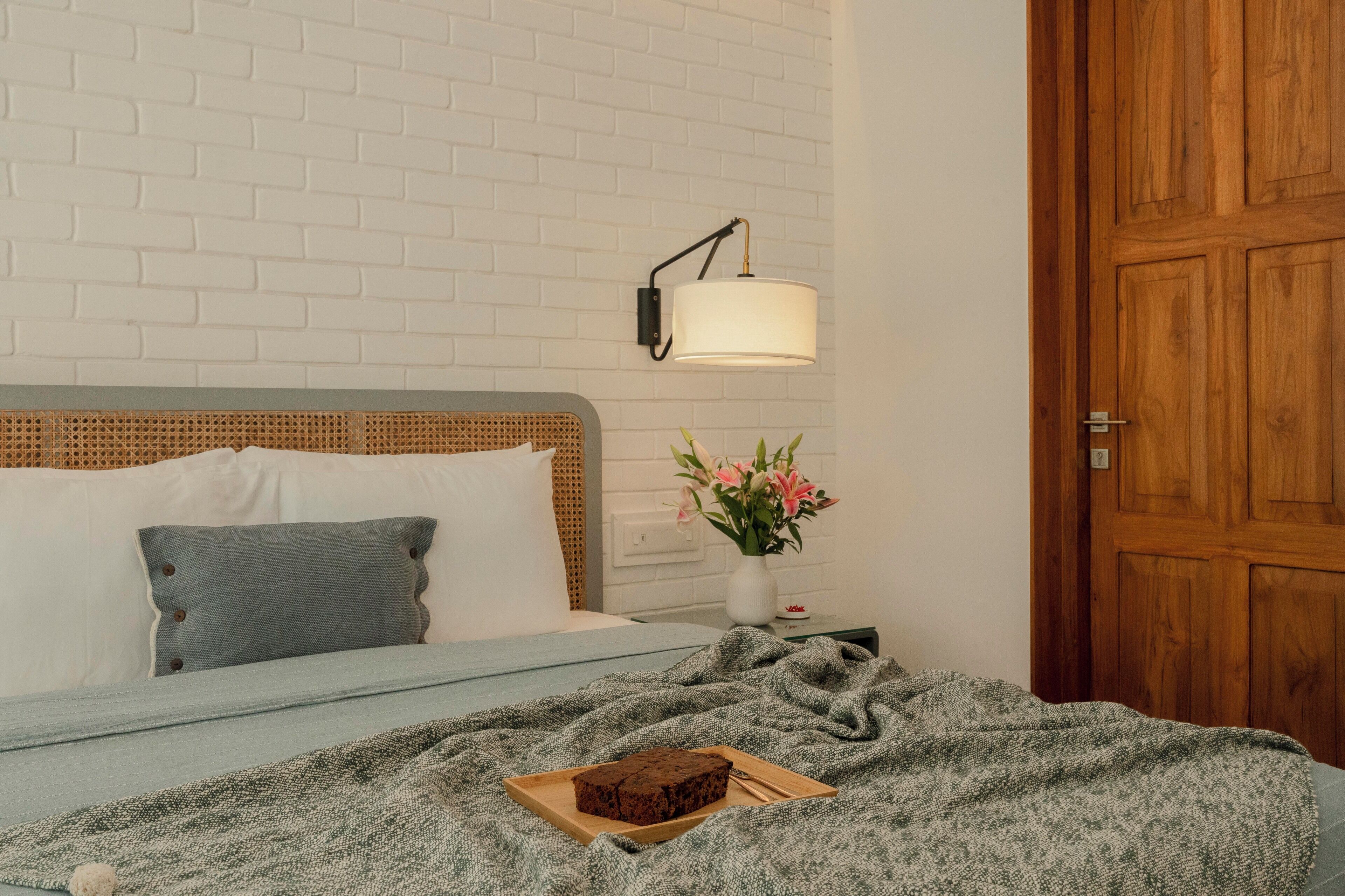 A bedroom with a bed, a bedside table with a lamp, a wooden door, and a small vase with flowers.