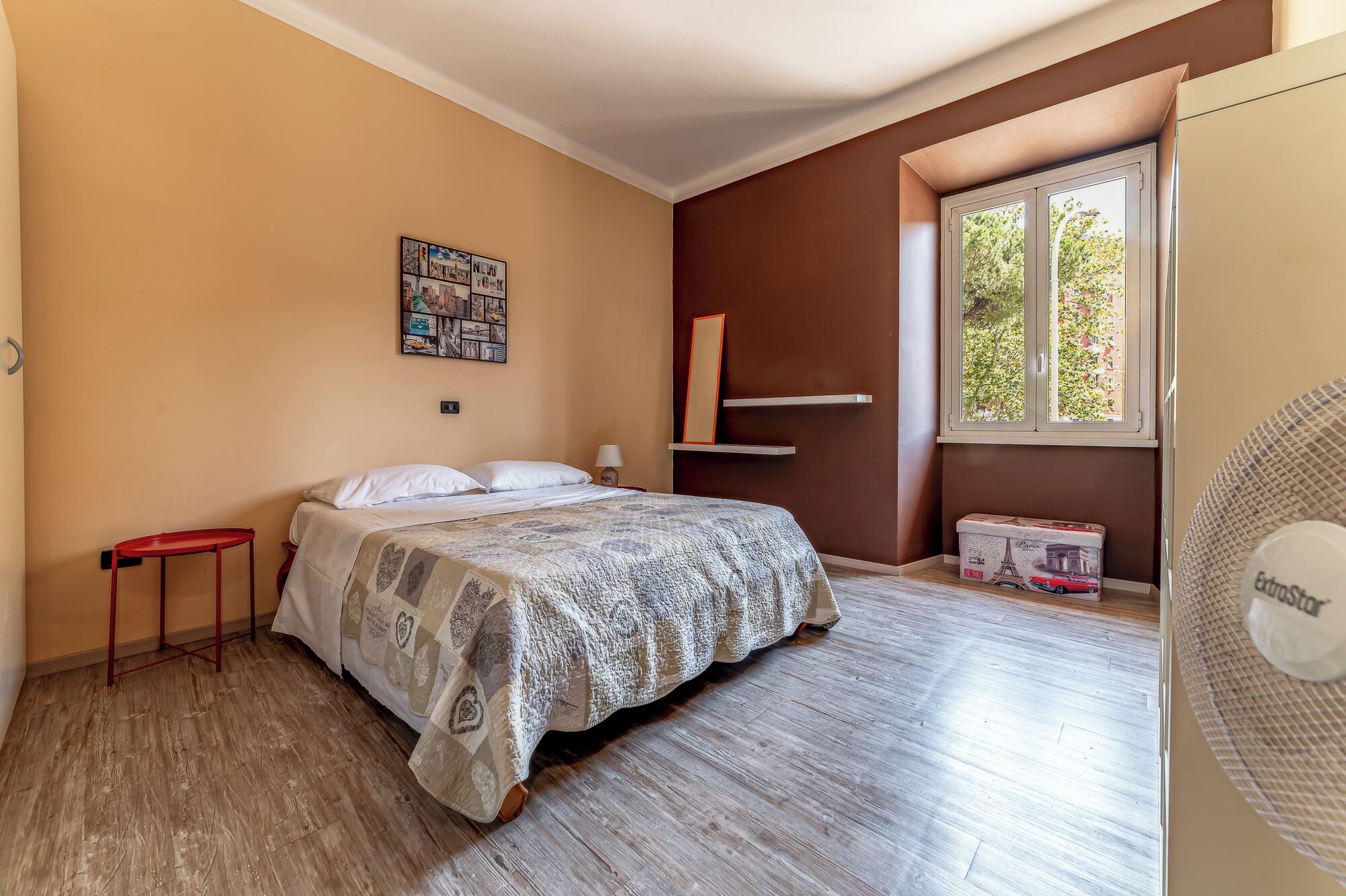 Un dormitorio con cama, una mesita, un espejo, una ventana con vistas y un ventilador.