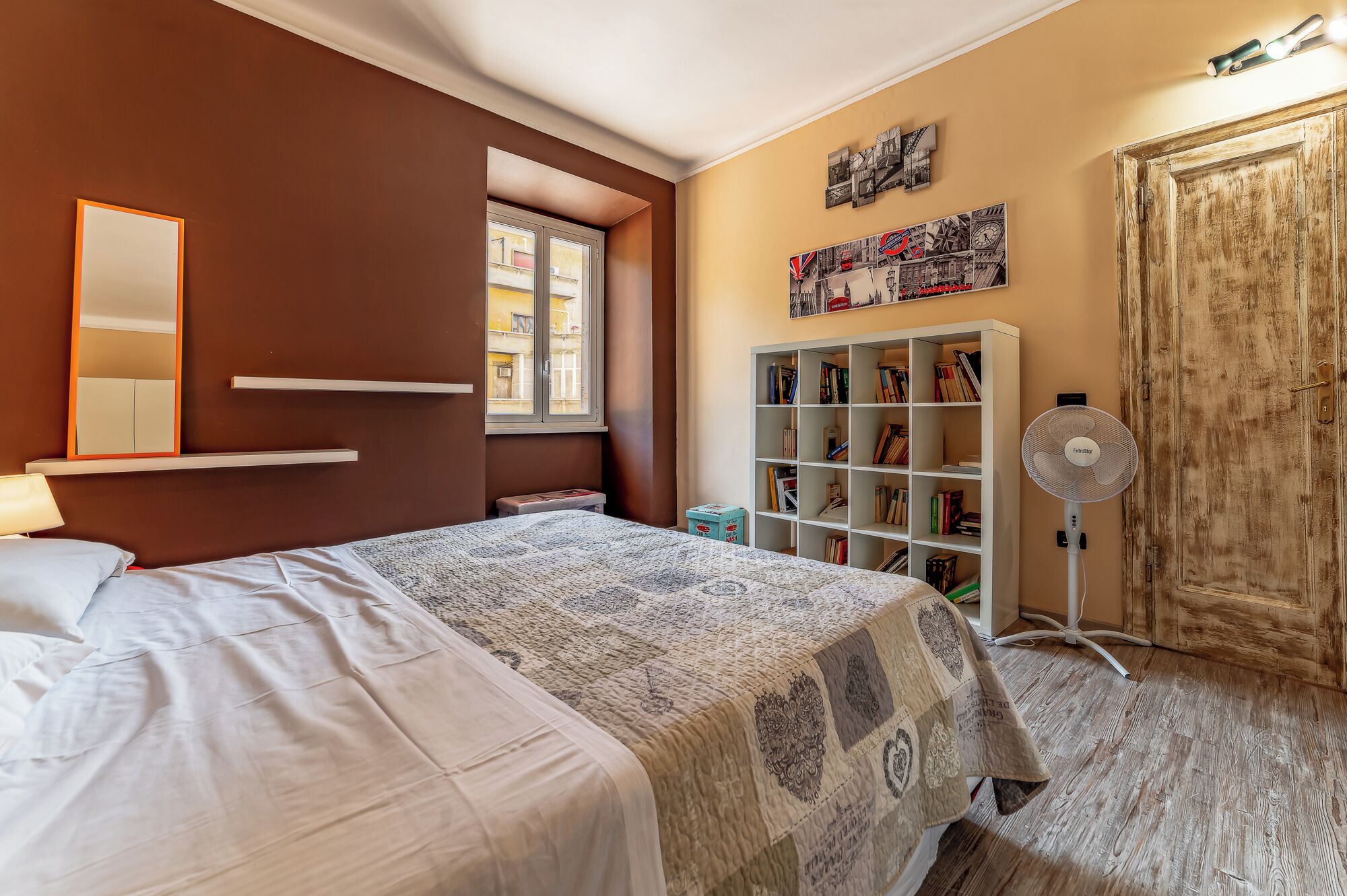 A bedroom with a bed, a bookshelf, a fan, and a door.