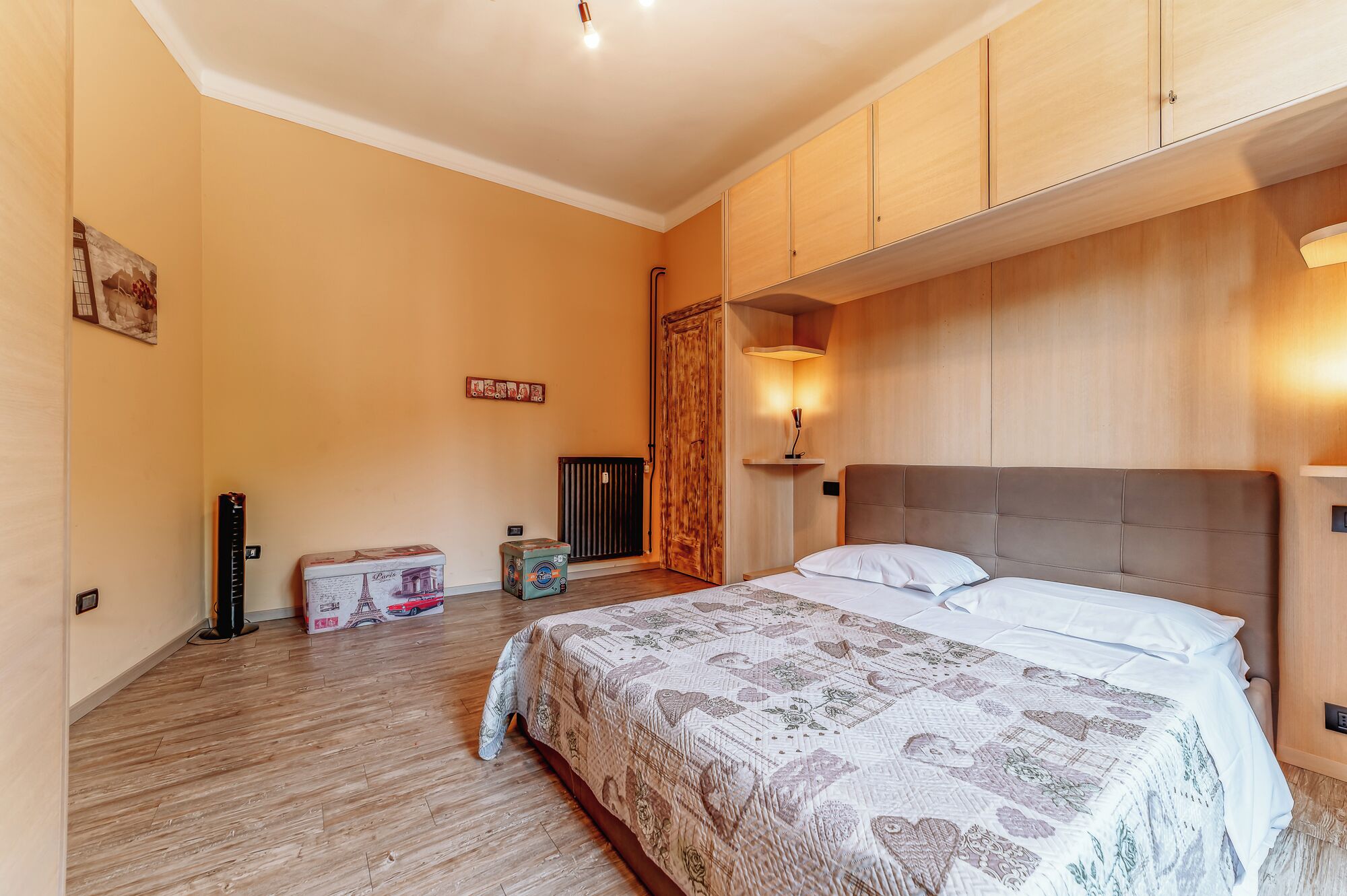 Une chambre à coucher comprenant un lit, une armoire en bois, un radiateur et une étagère fixée au mur avec une lampe.