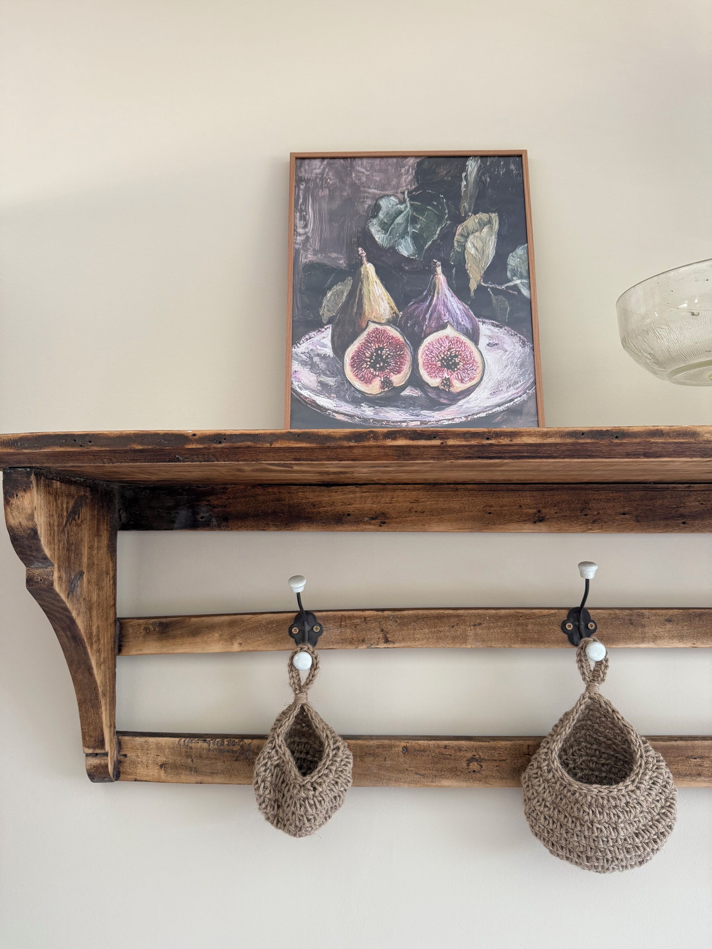 A wooden shelf with a framed painting of figs, two crocheted hanging items, and a clear glass object.