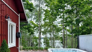 A wooden deck with a hot tub, a red building, and a forested area with a lake in the background.