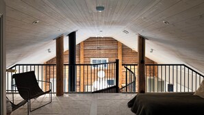 An attic bedroom with a sloped ceiling, a black metal railing, a black chair, a bed, and a view of the outside.