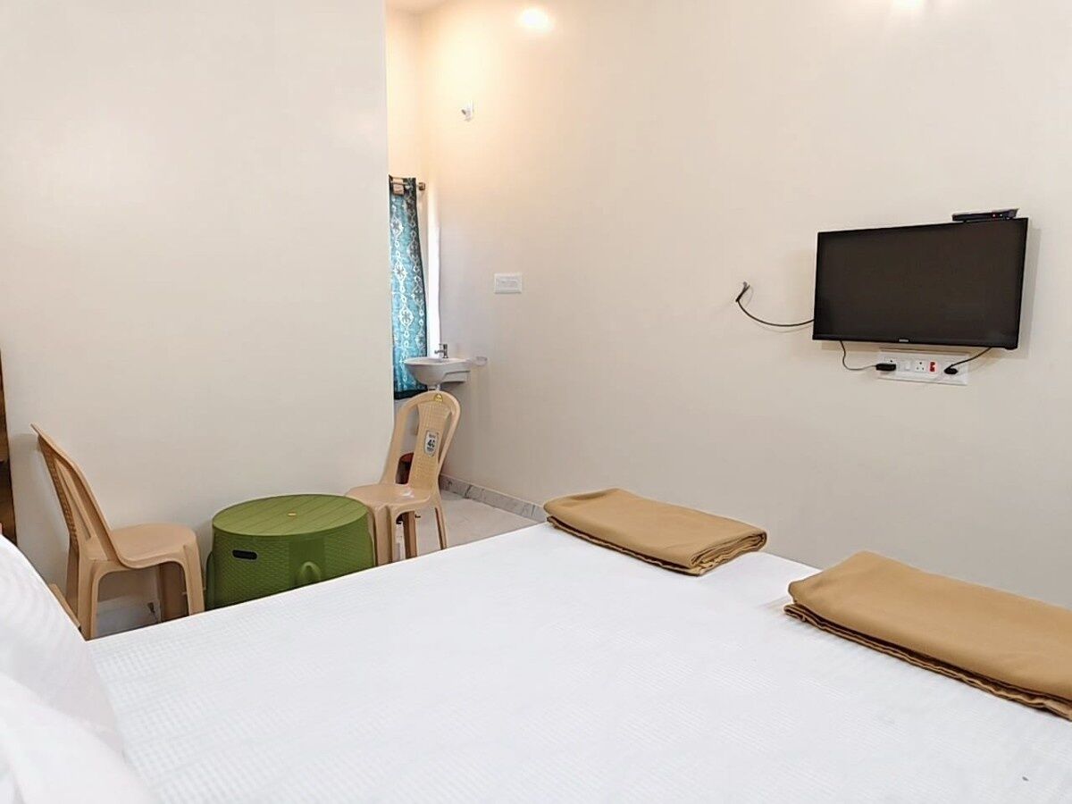 A hotel room with a bed, a television mounted on the wall, a small table with chairs, and a green stool.