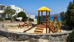 Ein Spielplatz mit einem gelben Spielhäuschen und Rutschen, umgeben von Steinmauern und Blick auf einen Strand.