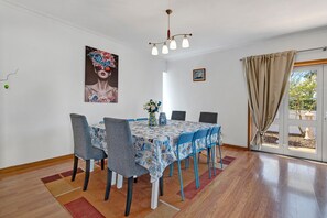 Un coin repas avec une table dressée pour six personnes, des chaises, un lustre, un tableau au mur et une vue à travers une porte vitrée.