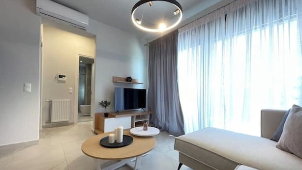 Een moderne woonkamer met een bank, een rond bijzettafeltje, een flatscreen televisie en een groot raam met gordijnen.