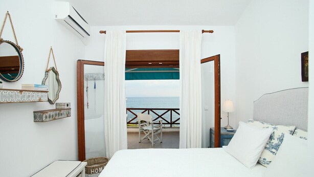 A hotel room with a bed, a view of the sea, a balcony with a chair, and a wooden ceiling. - Triton Maisonette by RentalsPro (Fourka)