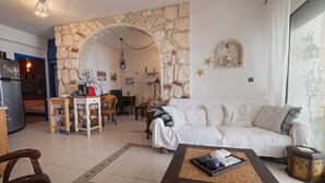 Ein Wohnzimmer mit Steinwand, einem weißen Sofa, einem Couchtisch mit Dekoration und einer Küchenzeile mit Kühlschrank und Esstisch.