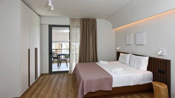 Habitación de hotel con cama, piso de madera, balcón con mesa y sillas, y vista al exterior.