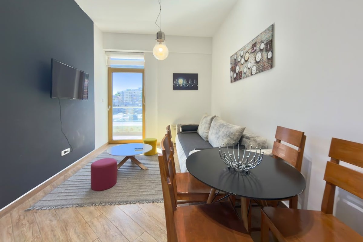 A modern living space with a dining area, a sofa, a TV mounted on the wall, and a large window.