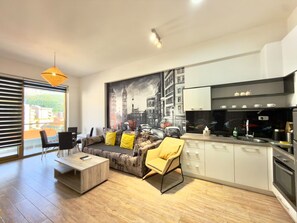 A modern living room with a sofa, coffee table, and a chair, a kitchen with a stove and cabinets, and a large window with blinds. - Orange Lux Apartments (Budva)