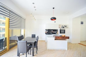 A modern kitchen with a central island, a dining area with a table and chairs, and a large window with blinds. - Orange Lux Apartments (Budva)