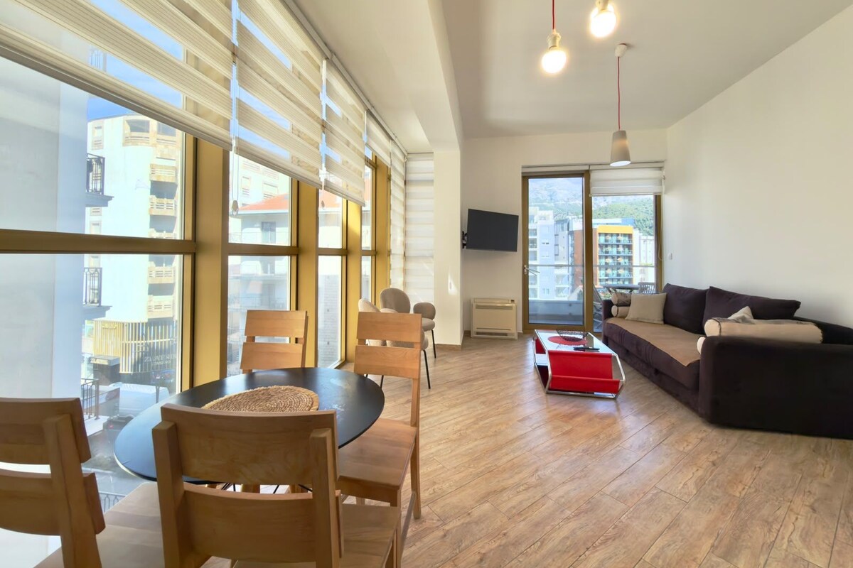 A modern living room with a round table, wooden chairs, a sofa, and a TV mounted on the wall.