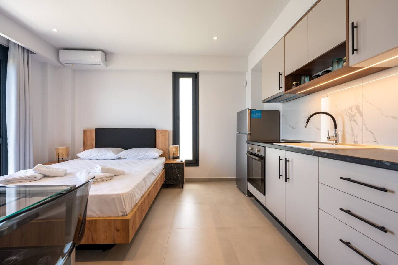 Moderni hotellihuone, jossa on sänky, yöpöydät, lasinen ruokapöytä, keittiöalue kaappeineen ja kodinkoneineen sekä suuri ikkuna, josta tulee runsaasti luonnonvaloa.