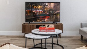 A living room with a TV displaying a cityscape, a wooden cabinet with board games, and two round tables. - SA Modern Home Nr River Walk + DT | BBQ Yard Games (San Antonio)