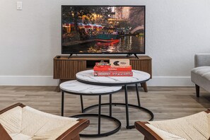 A living room with a TV displaying a cityscape, a wooden cabinet with board games, and two round tables. - SA Modern Home Nr River Walk + DT | BBQ Yard Games (San Antonio)
