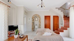 A living room with a sofa, a TV, a wooden table, and a staircase. - Azure Bay Villa by RentalsPro (Neos Marmaras)