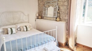 Una cama con estructura metálica blanca, una pared de piedra, un espejo con detalles decorativos y una ventana con cortinas.