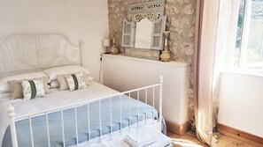 A bed with a white metal frame, a stone wall, a mirror with decorative elements, and a window with curtains. - This delightful two bedroom cottage offers accommodation for up to four people. (Salles-Lavalette)