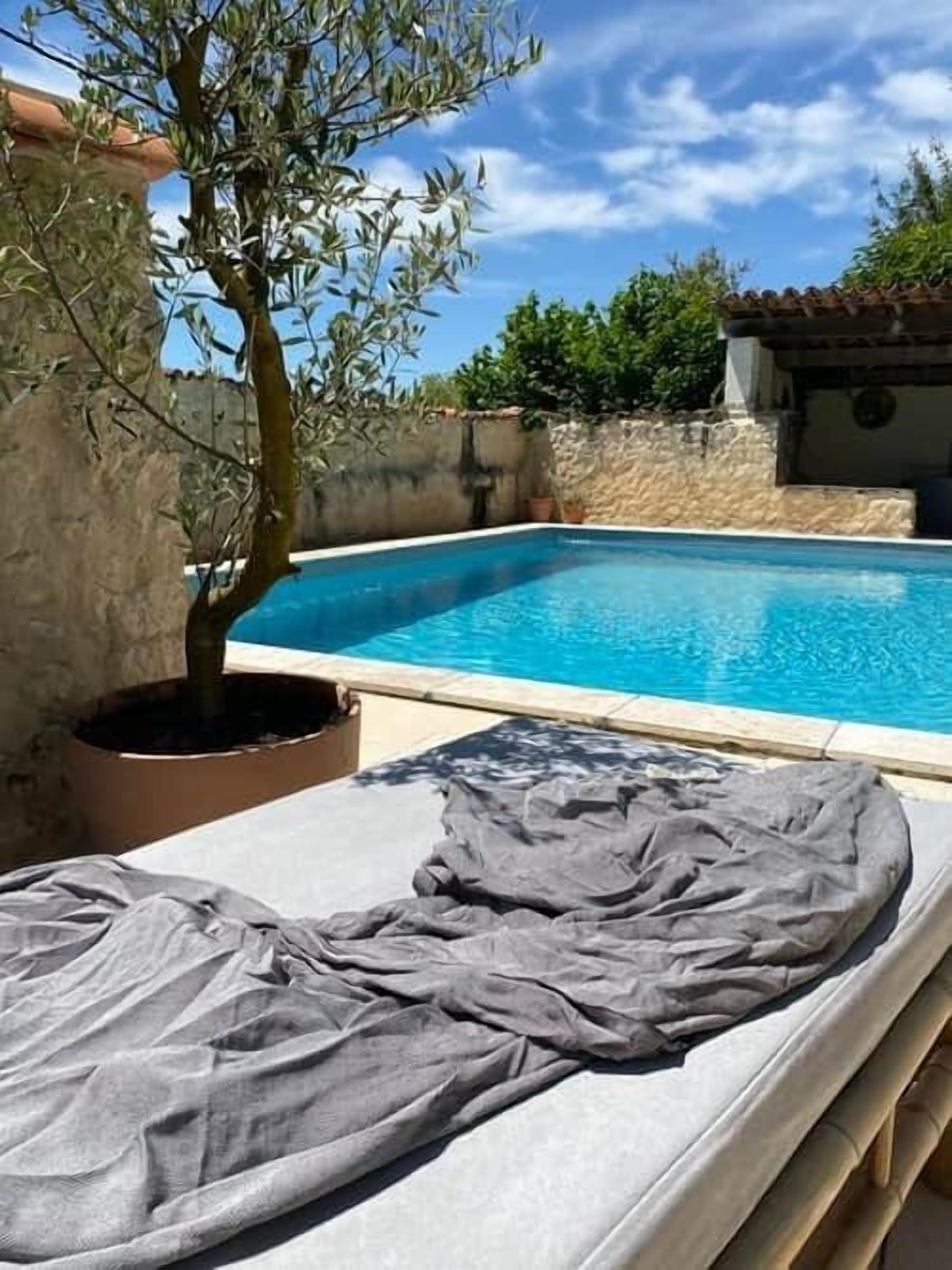 Zona junto a la piscina con una tumbona, un olivo en maceta y un muro de piedra.