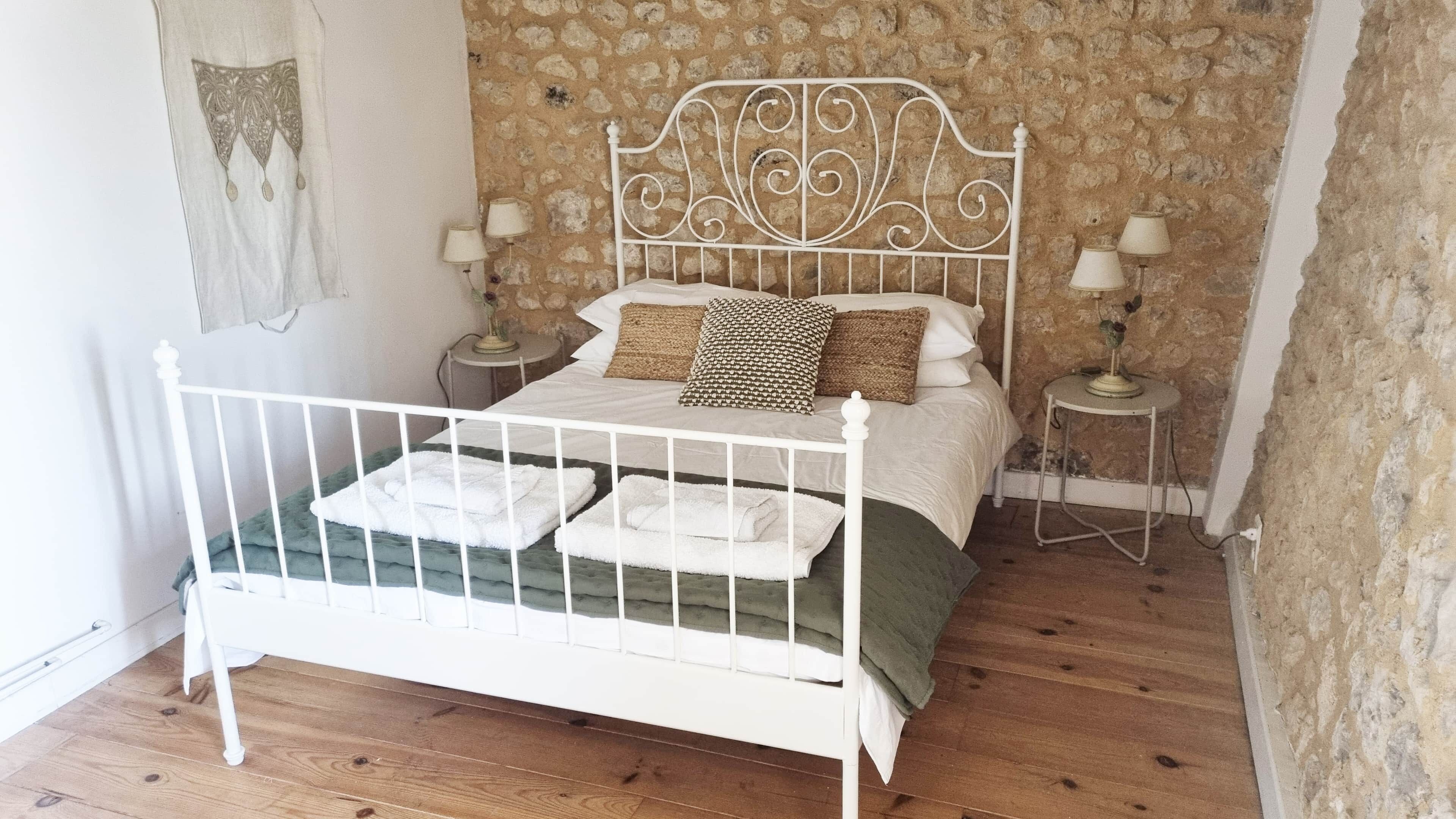 Una cama de metal blanco con una manta verde, dos mesitas de noche con lámparas y una pared con diseño de piedra.