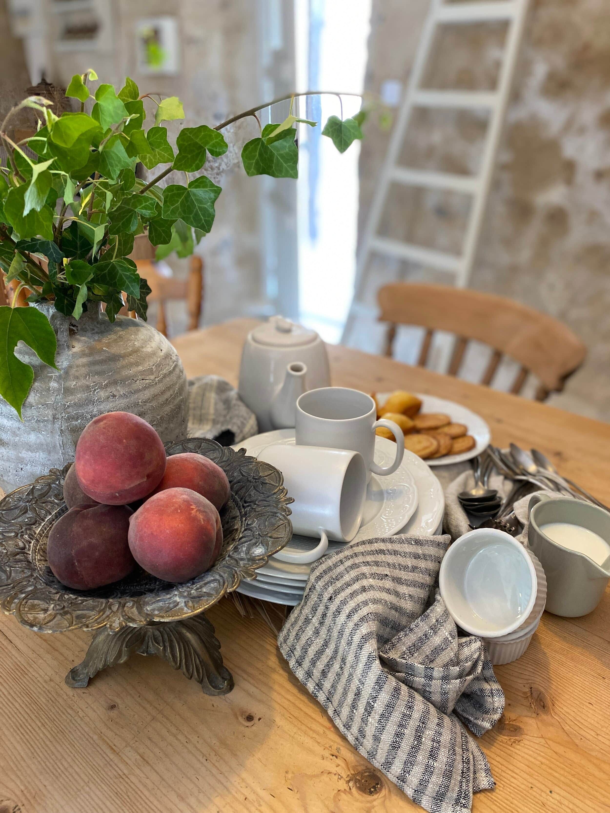 Une table en bois sur laquelle se trouvent un vase rempli de feuilles vertes, une assiette de fruits rouges, une théière, des tasses et un bol de lait.