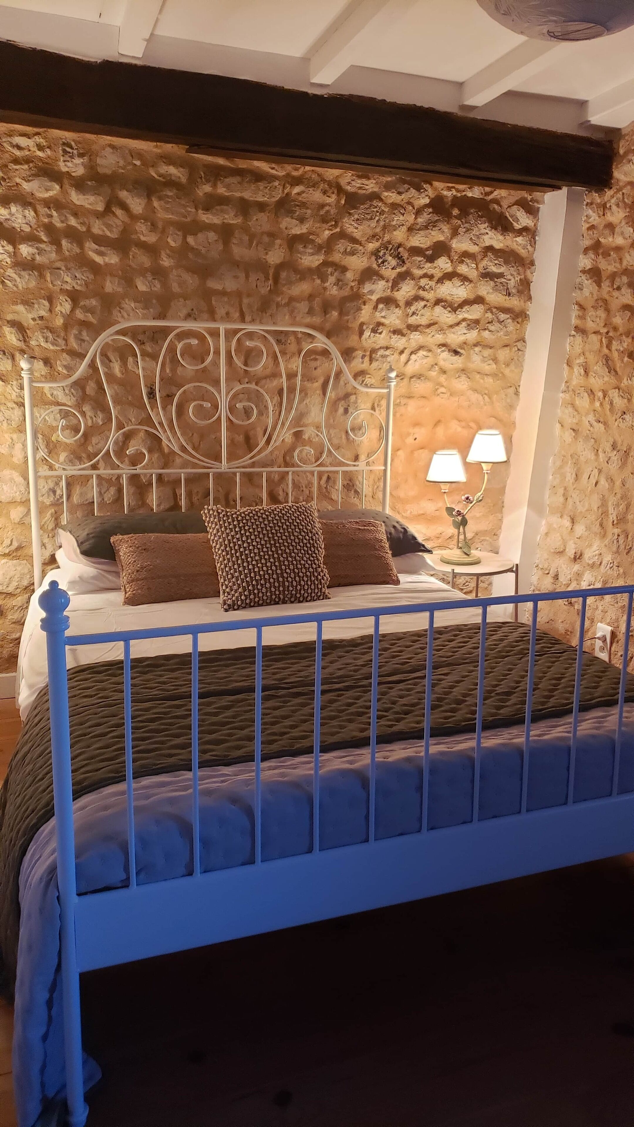 A bedroom with a stone wall, a metal bed frame, a bedside table with a lamp, and a bed with a patterned blanket and pillows.