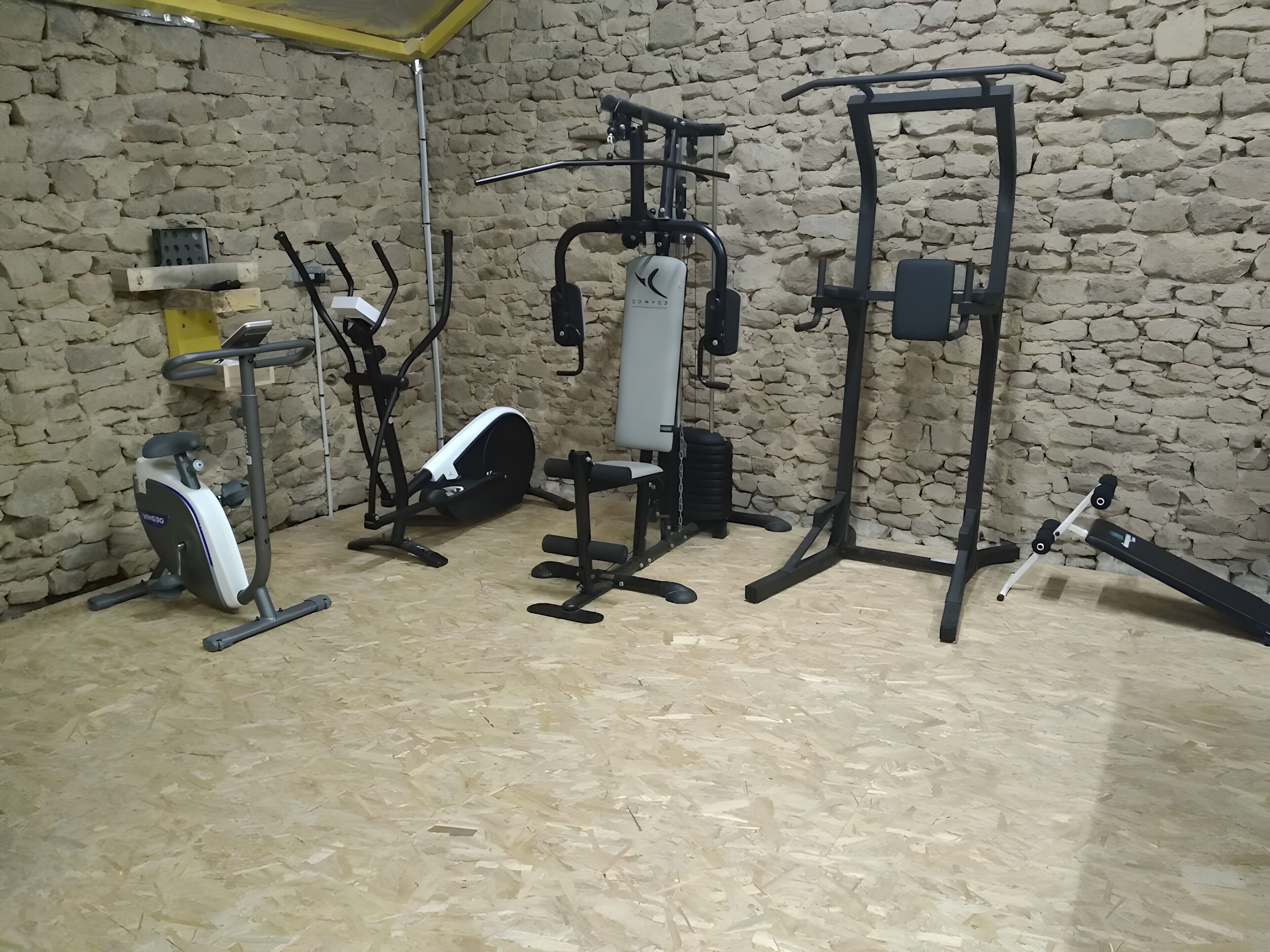 Een sportschool met diverse fitnessapparaten, waaronder een hometrainer, een spinningfiets en een krachttrainingapparaat, met een achtergrond van een stenen muur.