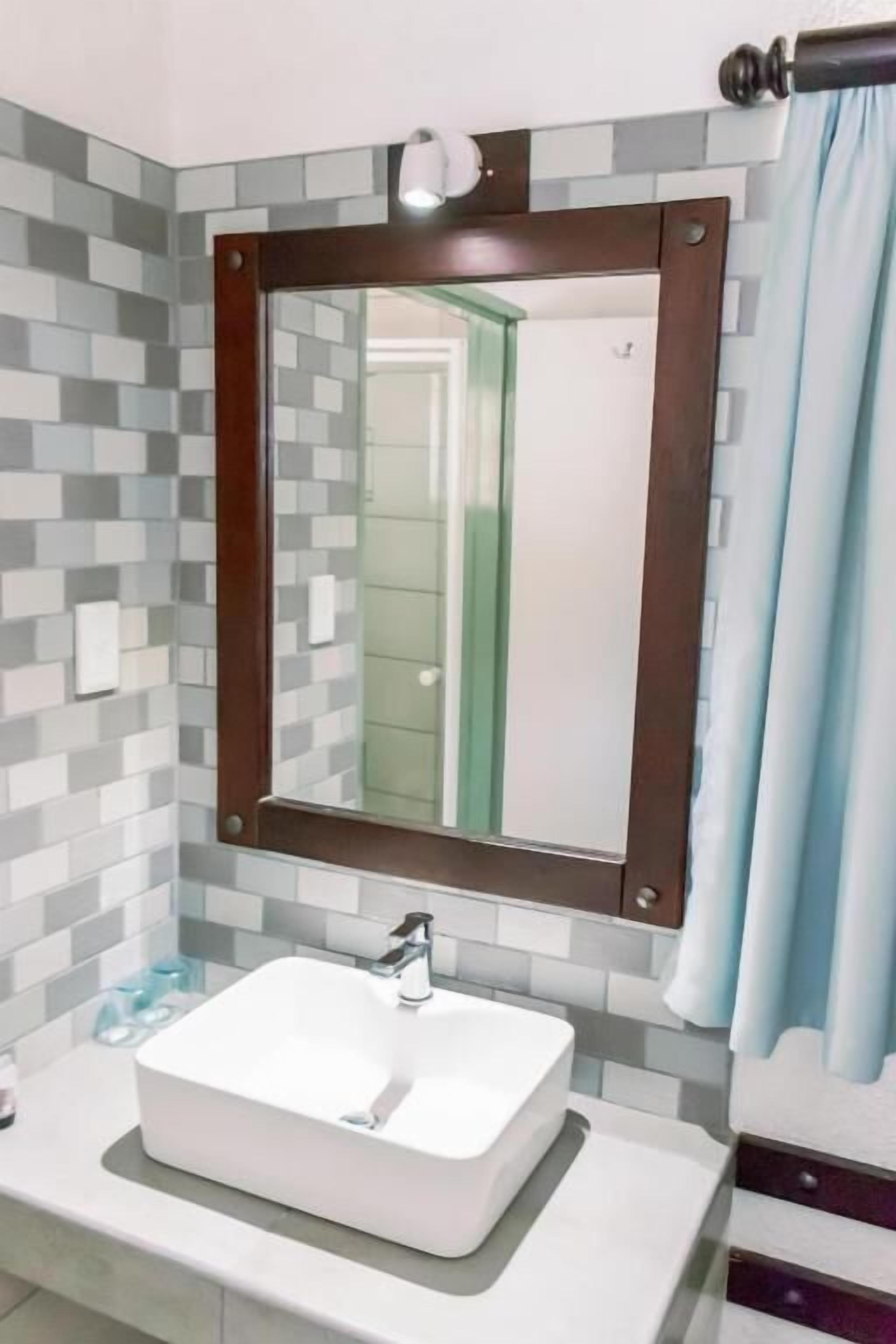 Une salle de bain avec un lavabo blanc, un miroir avec un cadre en bois et un mur recouvert de carreaux.