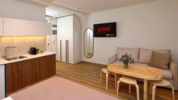 Een moderne woonkamer met een houten eettafel, een bank, een keuken met een spoelbak en een aan de muur bevestigde televisie waarop 'NETFLIX' wordt getoond.