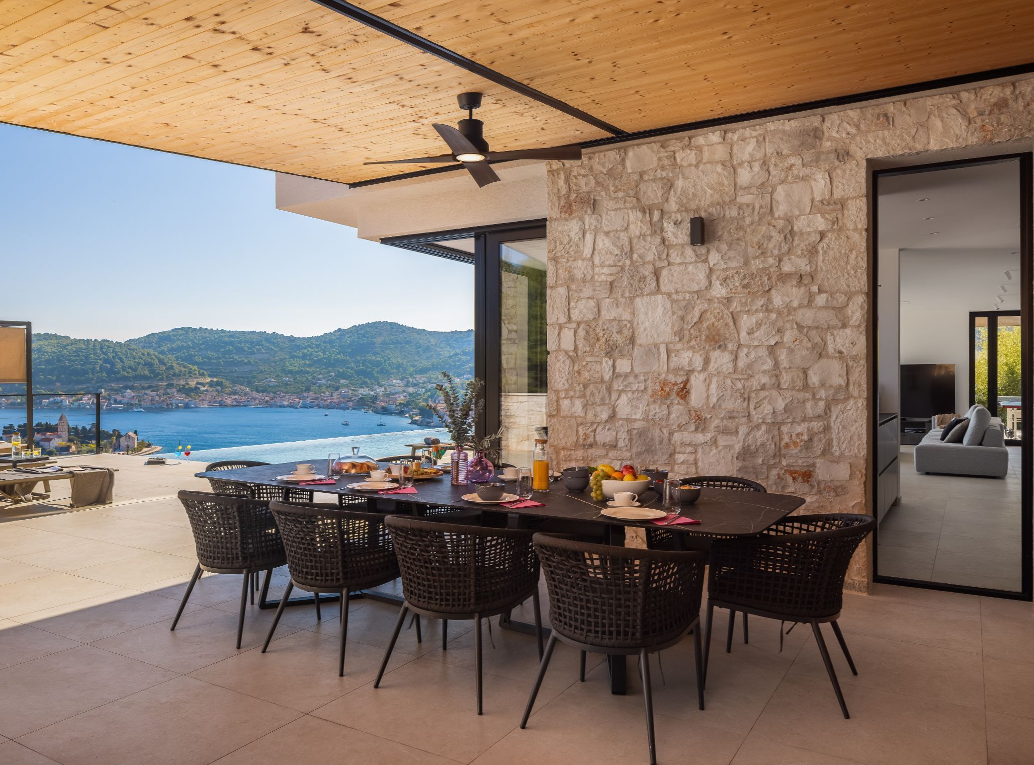 A dining area with a table set for four, a ceiling fan, stone walls, and a view of the sea.