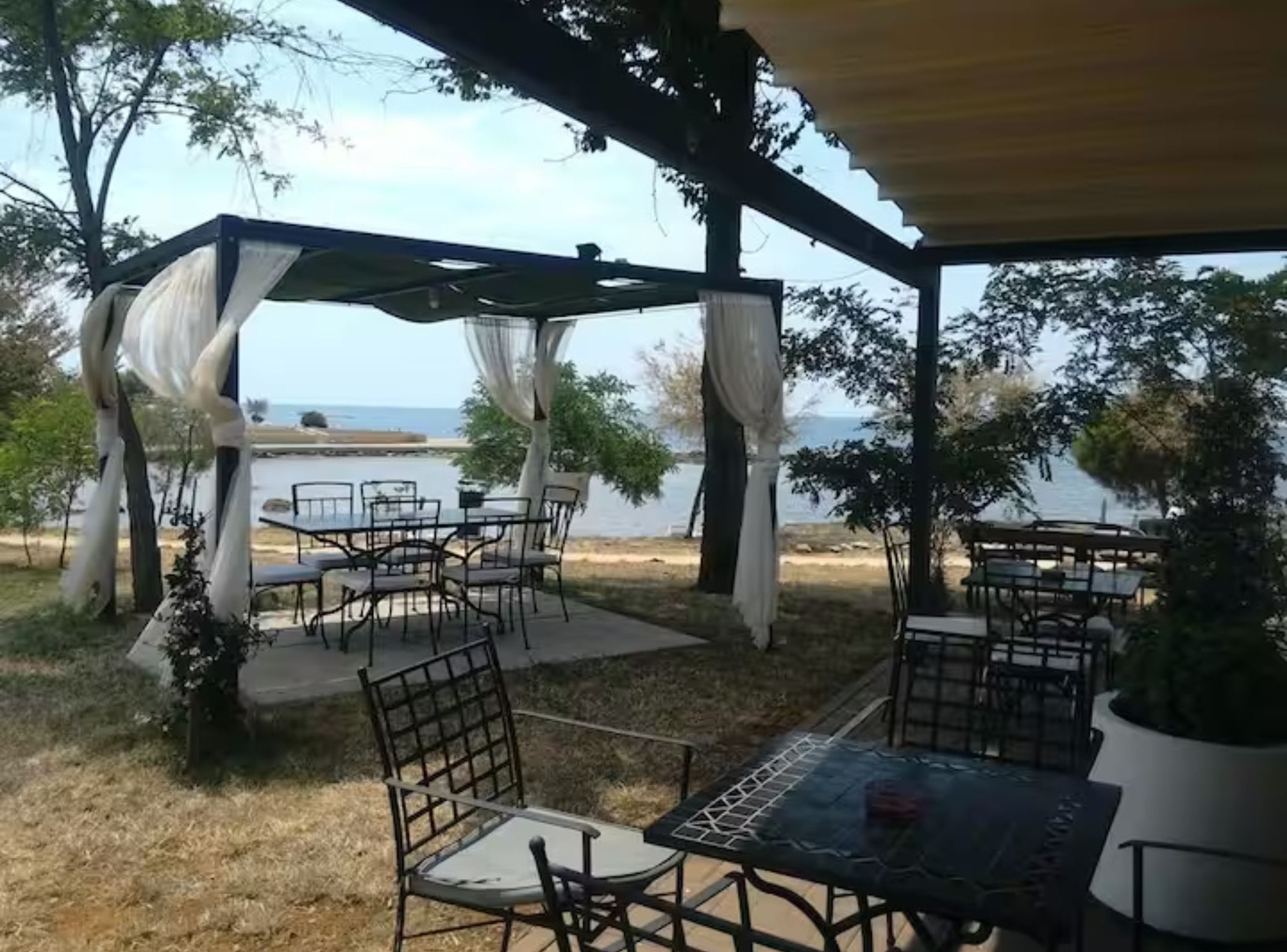 An outdoor patio with tables and chairs, a pergola with draped fabric, and a view of the sea.