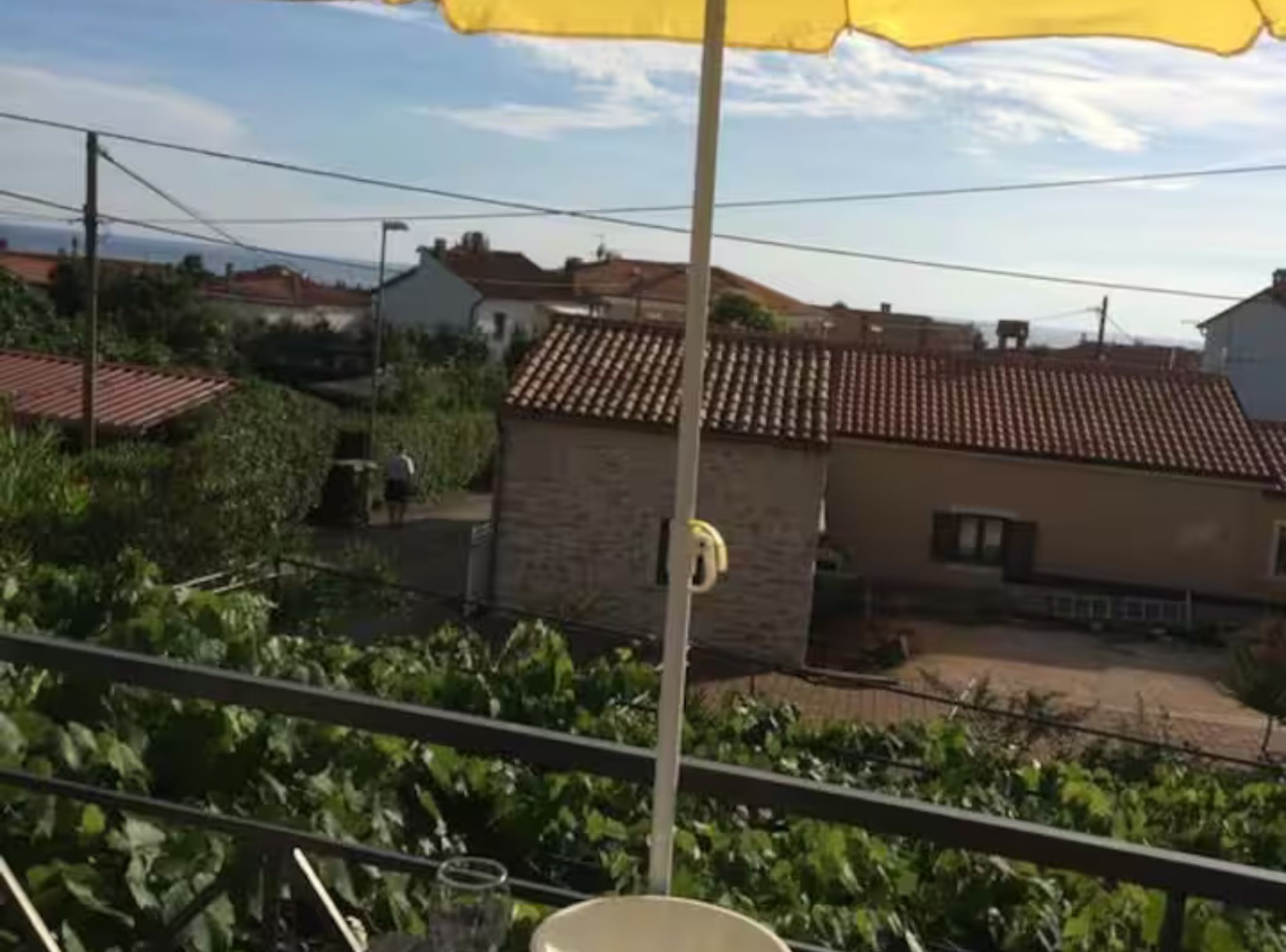 A balcony with a yellow umbrella, a glass of water, and a view of houses with red-tiled roofs.