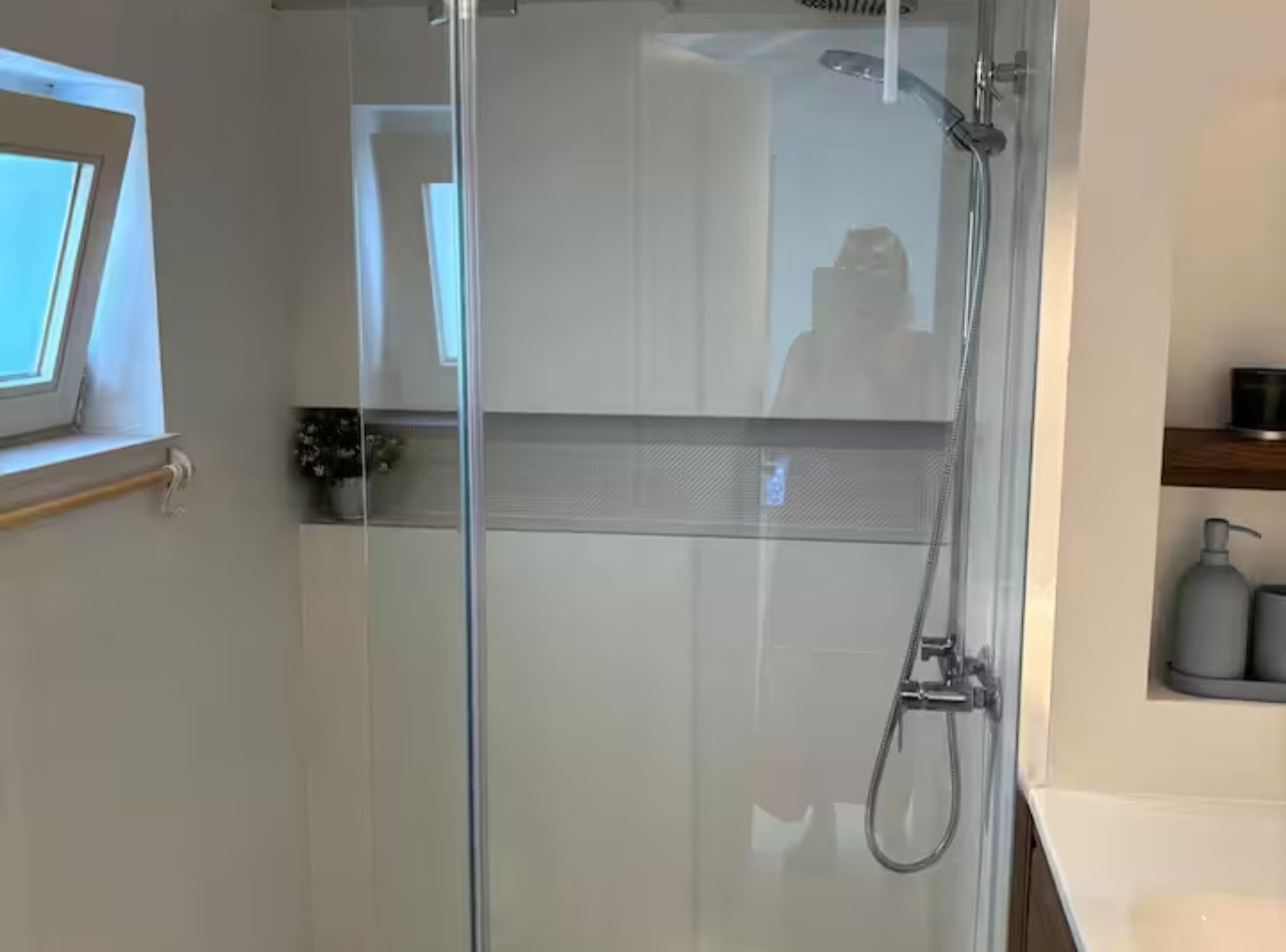 A glass-enclosed shower with a handheld showerhead, a window, and a small shelf with toiletries.