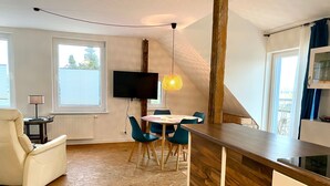 Eine moderne Küche mit Holzarbeitsplatte, ein Essbereich mit rundem Tisch und Stühlen, ein an der Wand montierter Fernseher und ein Fenster mit Jalousien.