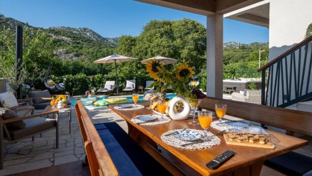 An outdoor dining area with a view of the mountains, wooden furniture, and a table set with a vase of sunflowers, fruit, and drinks.