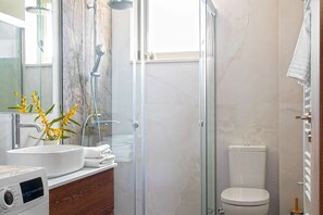 A modern bathroom with a glass shower enclosure, a toilet, and a sink with a mirror and flowers. - Vineyard View, next to Best Wineries, "Trigos" appartment (52QJ+W6 Avgeniki)